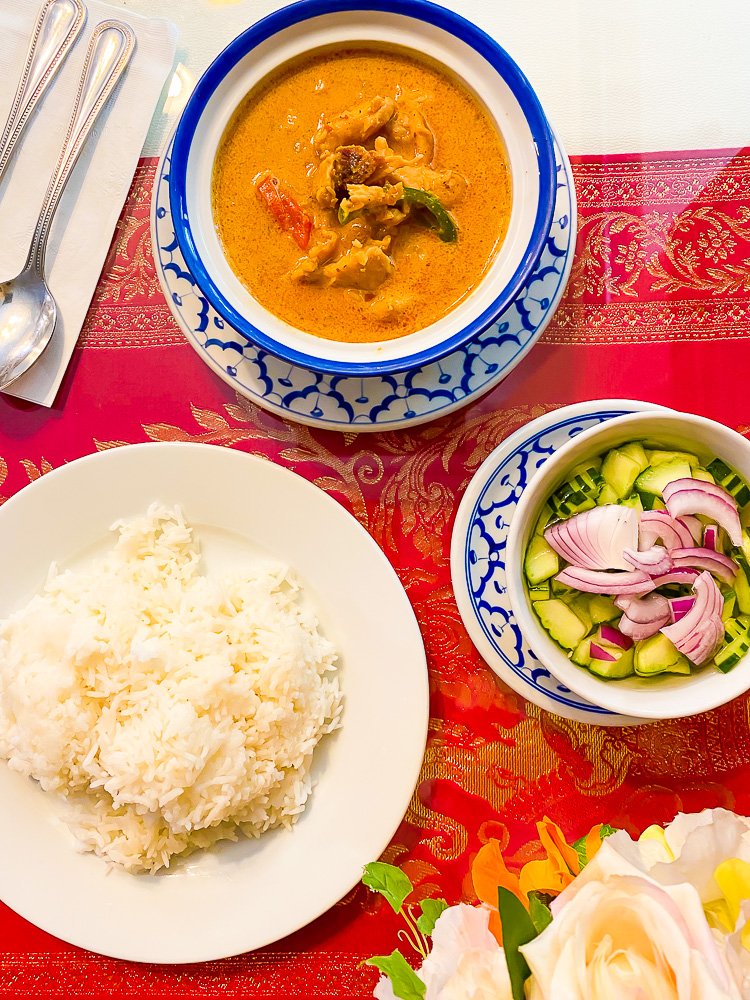 bowl of panang curry, plate of rice, and bowl of Thai cucumber salad on red tablecloth.