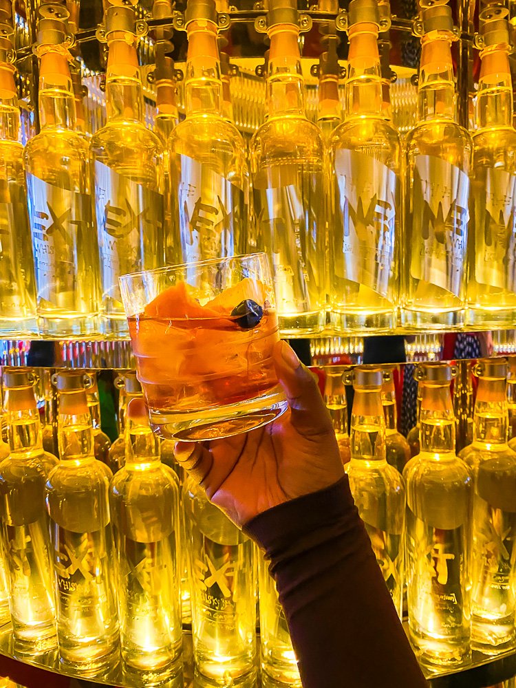 holding old fashioned cocktail with orange peel and cocktail cherry garnish in front of display of Japanese whiskey bottles.