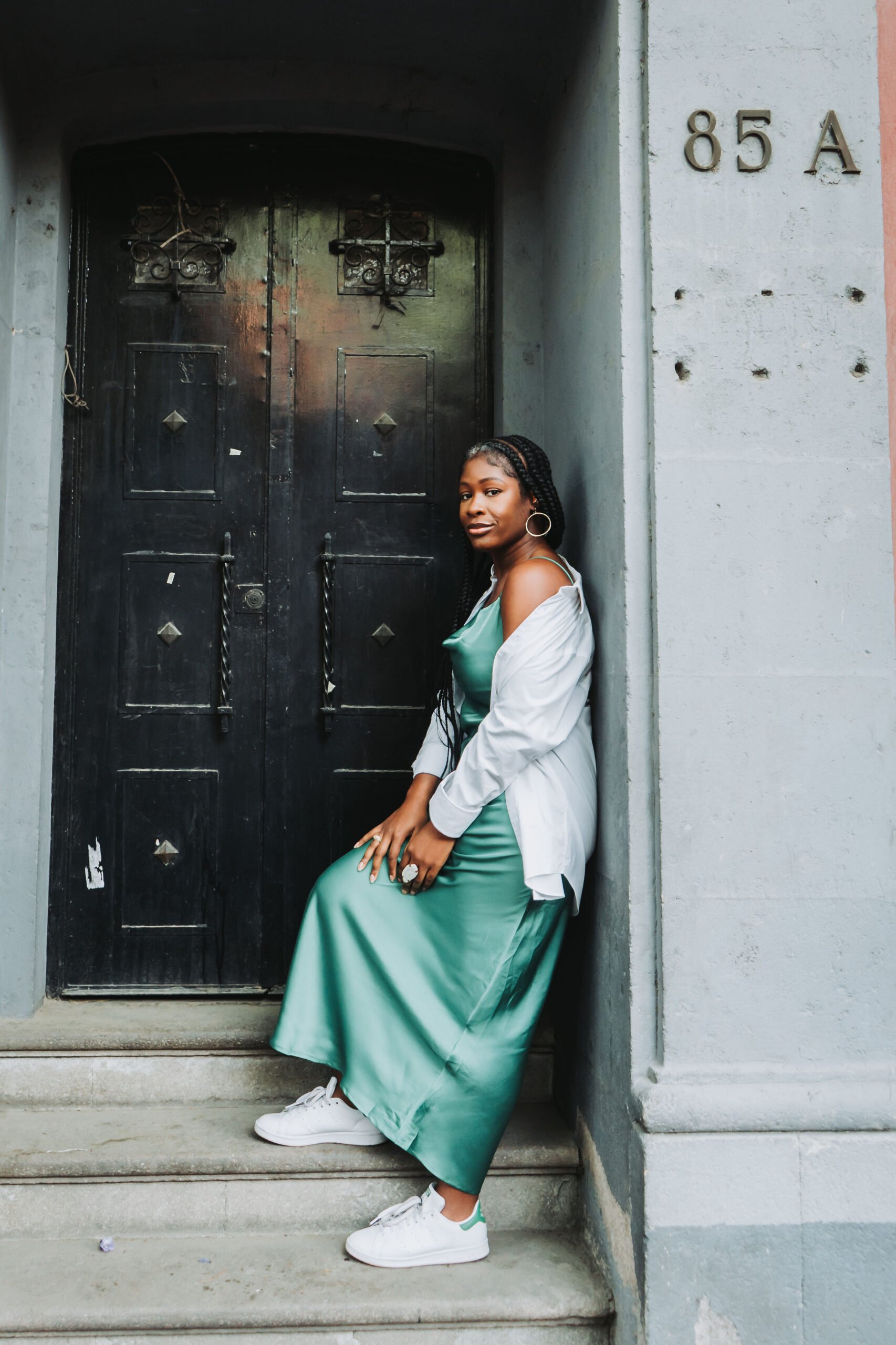 Jazzmine standing on the steps of a porch in Coyoacán, Mexico City, wearing a long green silk dress, white oversized button down shirt, white adidas sneakers, and white stone ring.