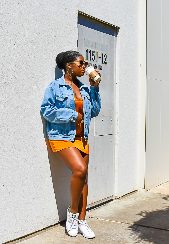 Jazzmine leaned against wall wearing orange slip dress, oversized denim jacket, and stan smith sneakers.