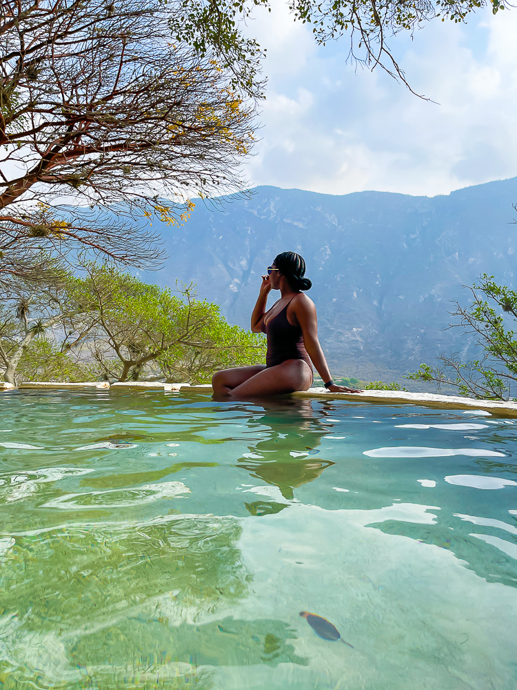 Jazzmine perched on ledge of a Tolantongo gruta hot spring pool in a chocolate one piece bathing suit looking at view of trees and a distant mountain.