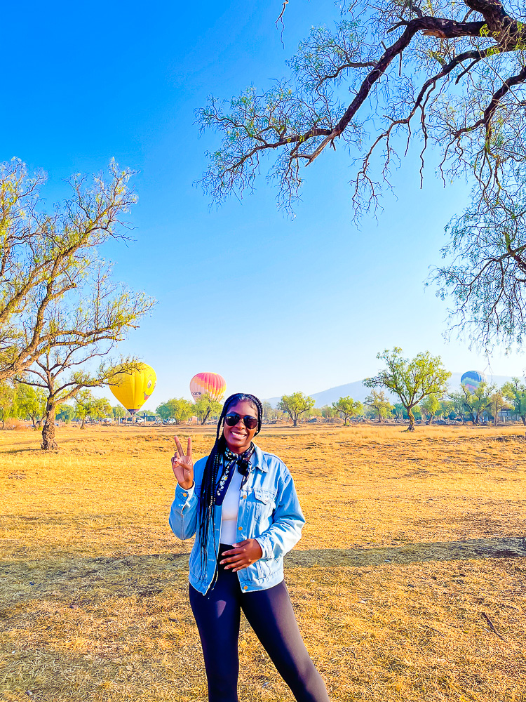 Jazzmine posing in front of hot air balloons landing in a grassy field.