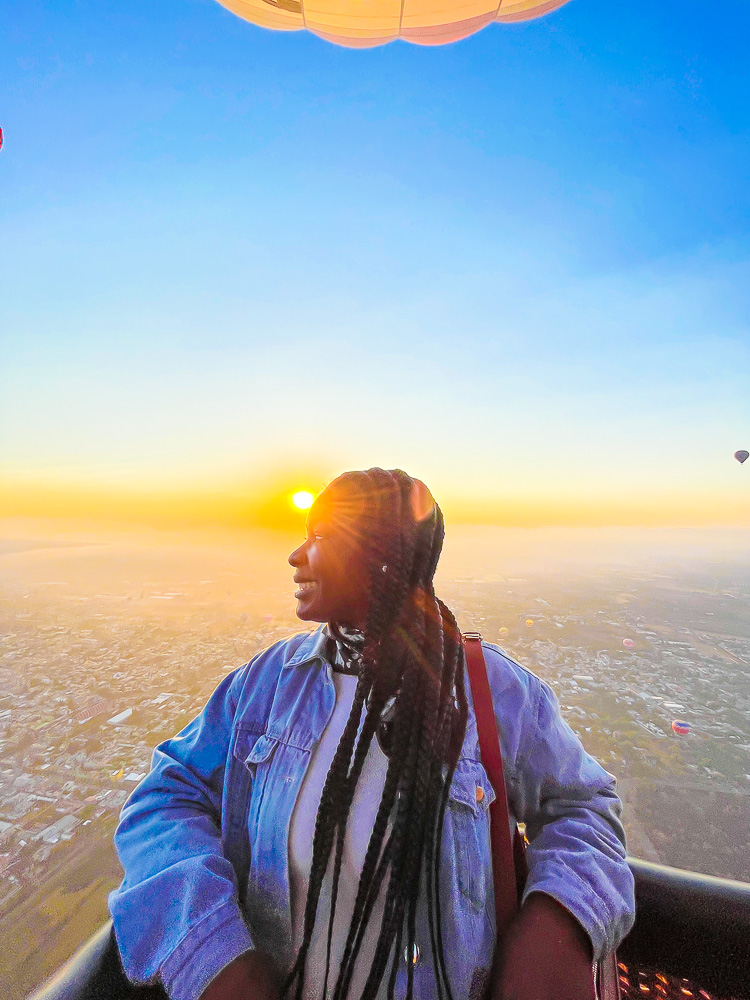 Jazzmine leaning against side of a flying hot air balloon gondola in Mexico taking in the view as the sun rises behind her.