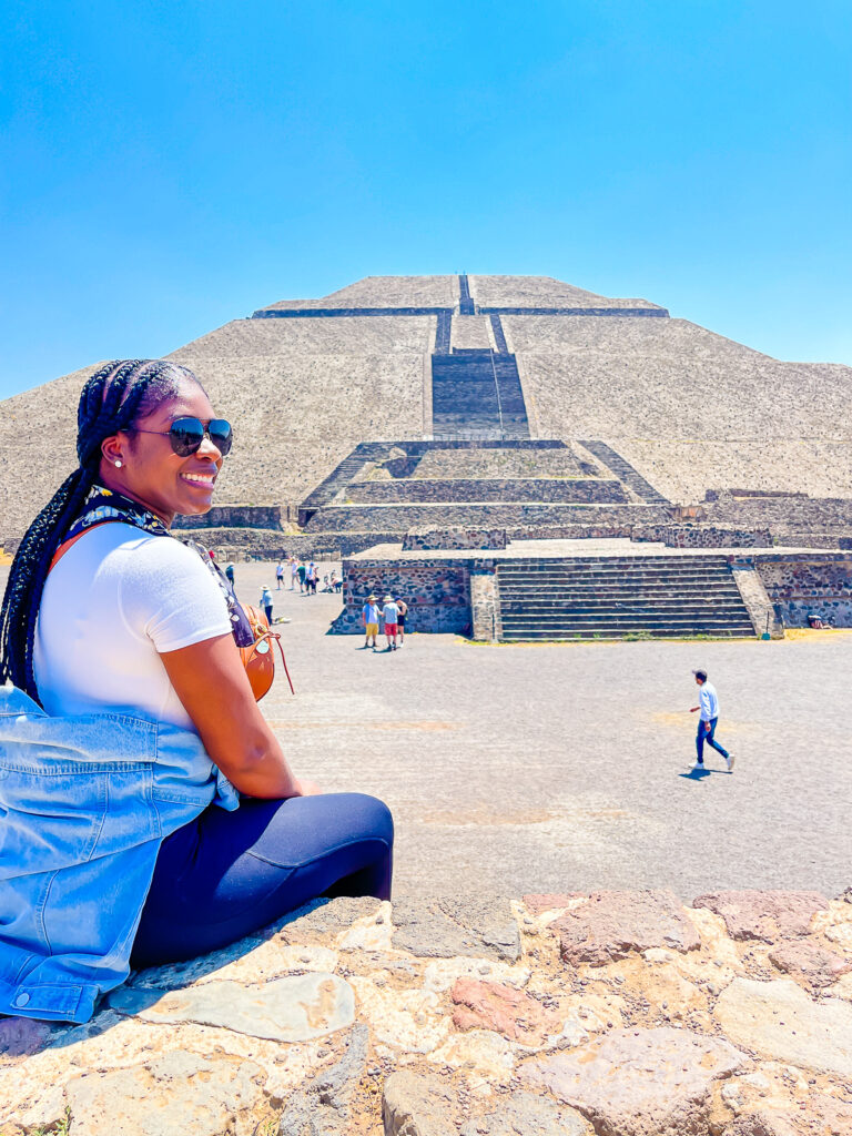 Jazzmine wearing black aviator sunglasses smiling and perched on a ledge looking out at Pyramid of the Moon in Teotihuacan archaeological zone.
