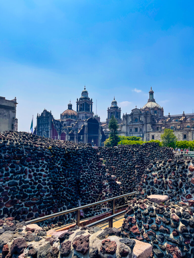 Stone wall of Templo Mayor ruins set against the backdrop of Spanish churches in Mexico City's historic central district.