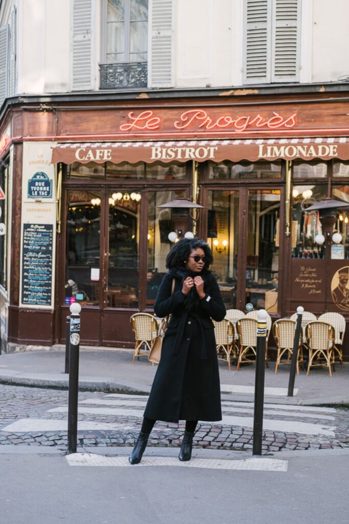 Jazzmine standing in street of Montmartre neighborhood of Paris, France clutching the fur collar of a long black coat.