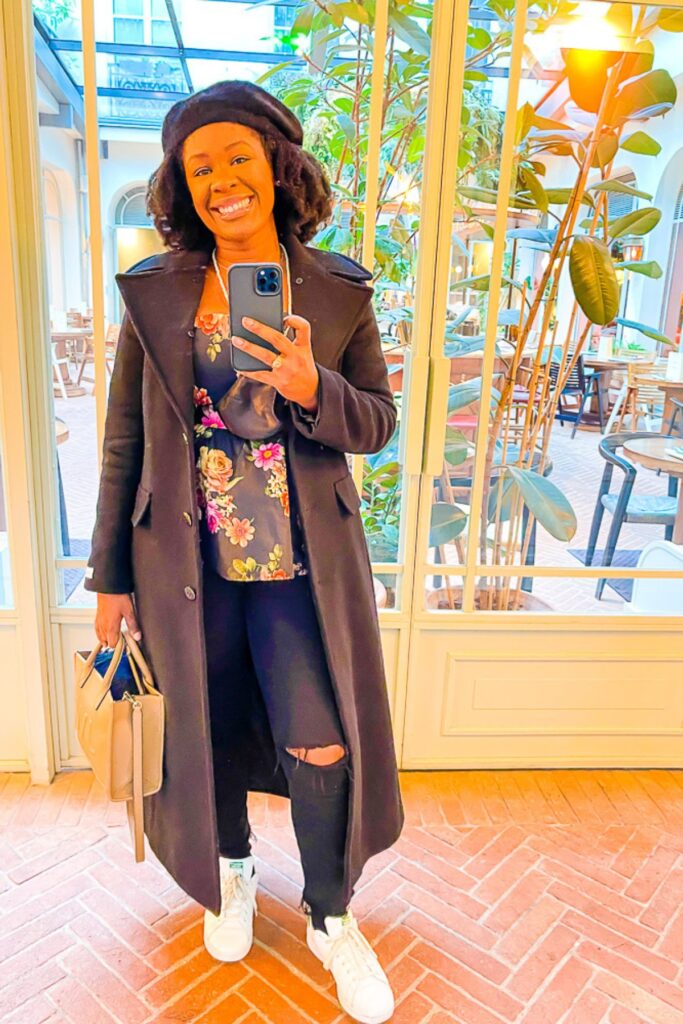 Jazzmine taking mirror selfie at Le Grands Boulevards Hotel wearing long black coat, floral peplum blouse, black jeans, sneakers, and a beret.