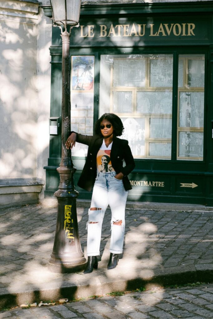 Jazzmine posed against streetlight in front of French storefront wearing a graphic t shirt, double breasted black blazer, and ripped jeans.