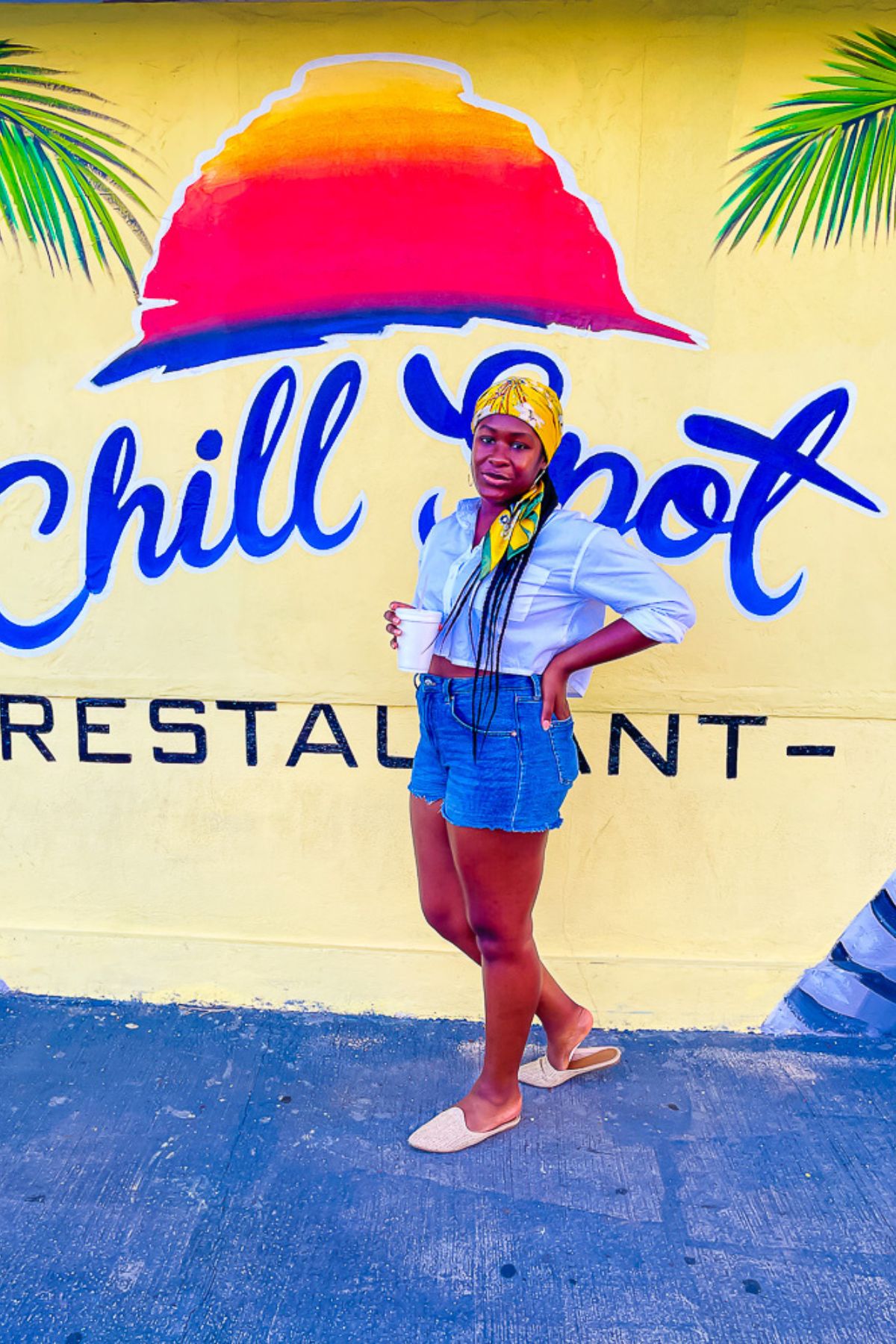 Jazzmine standing outside the Cayman Islands Chill Spot restuarant wearing a white top, denim shorts, and colorful scarf on her head.