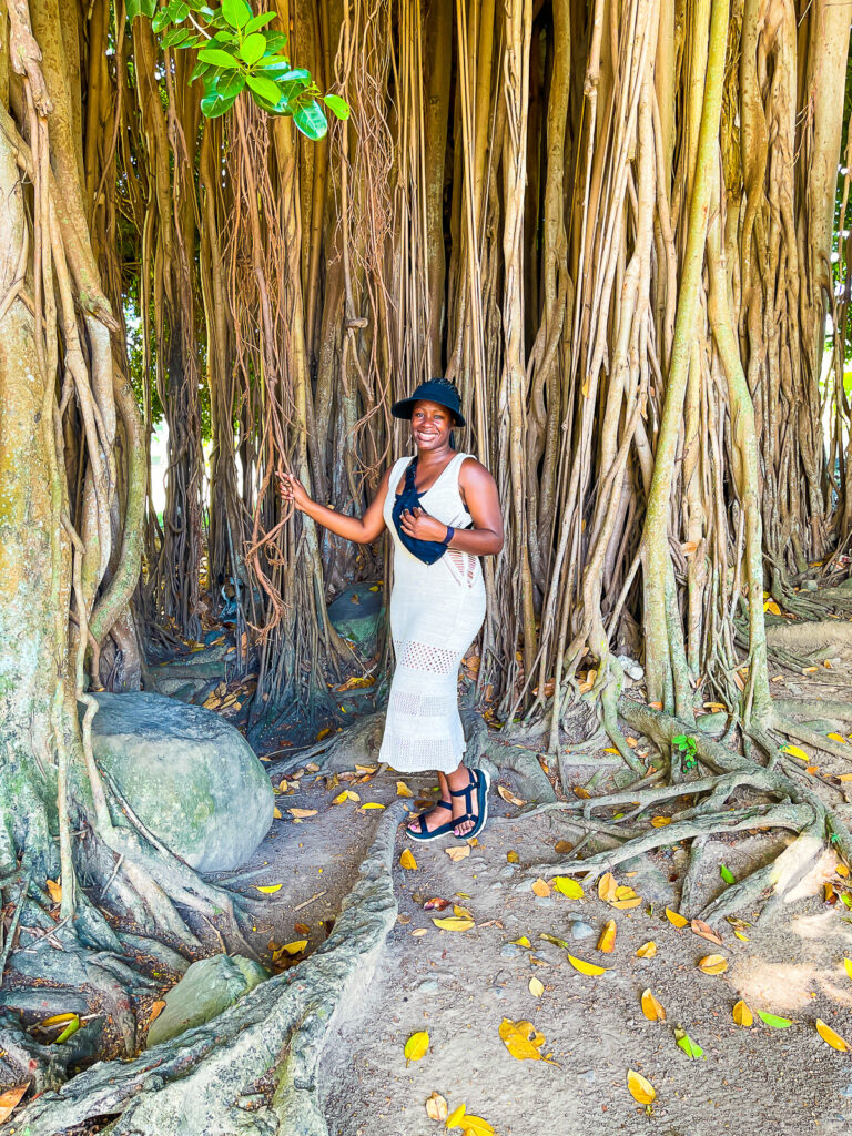 Jazzmine standing inside massive banyan tree outside Dominica's botanic gardens in Roseau; she's wearing a crocheted dress, black bum bag, black Tevas sandals, and. a black visor.