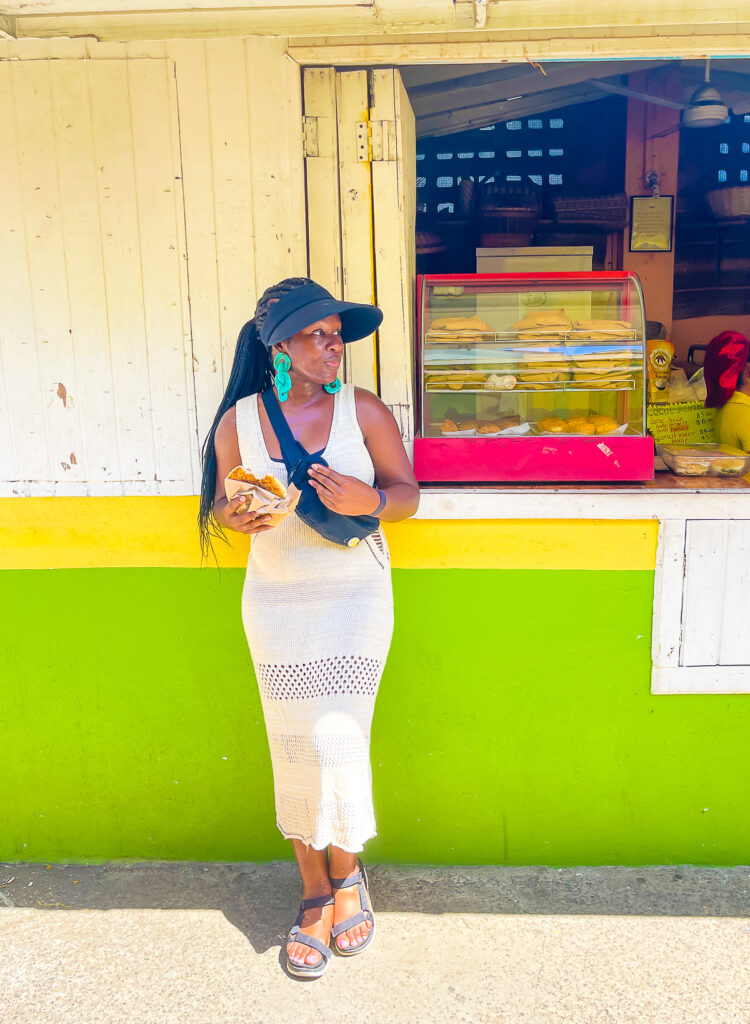 Jazzmine standing in front of bakery stall at Roseau Old Market in Dominica; wearing a tan crochet dress, black sandals, black hat, and turquoise earrings.