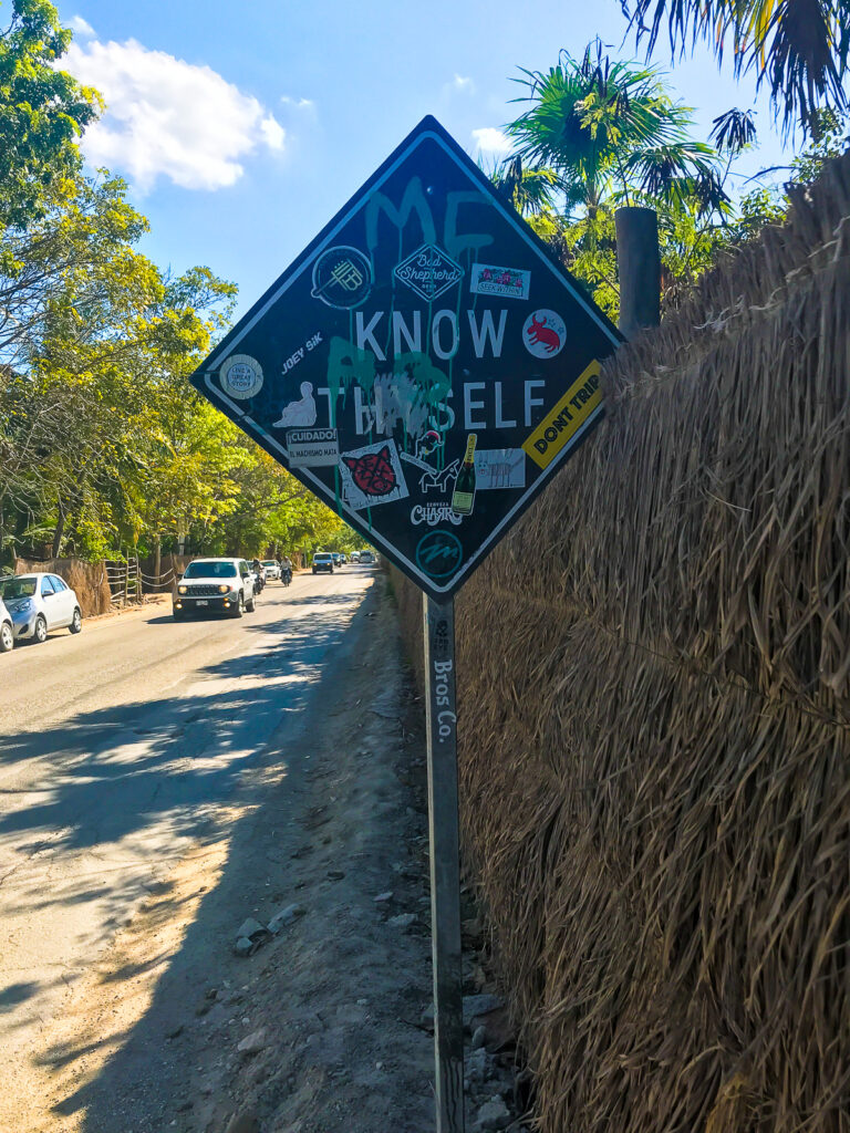 Black and white sign that reads "know thyself" on roadside in Tulum, Mexico, with cars and motorcyles driving by.