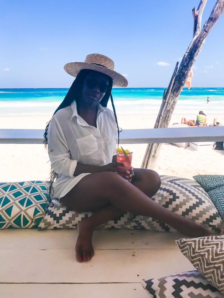 Jazzmine seated on colorful patterned floor cushions at Coco Beach Tulum; holding a pink pulpo cocktail with the blue ocean and sand behind her.