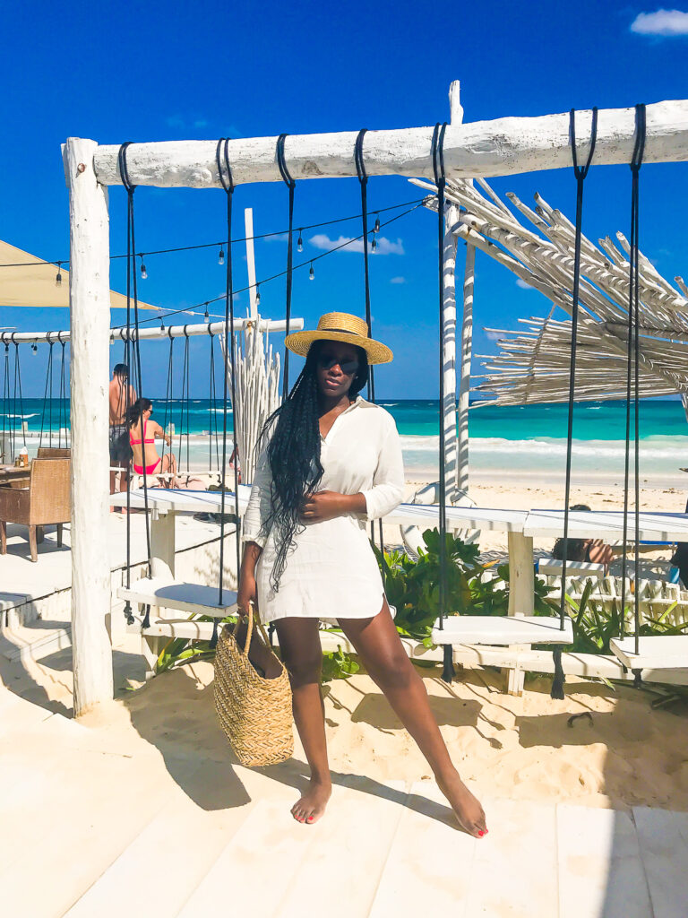 Jazzmine posing in front of swings at Coco Beach Club in Tulum wearing a white cover-up dress and straw panama hat.