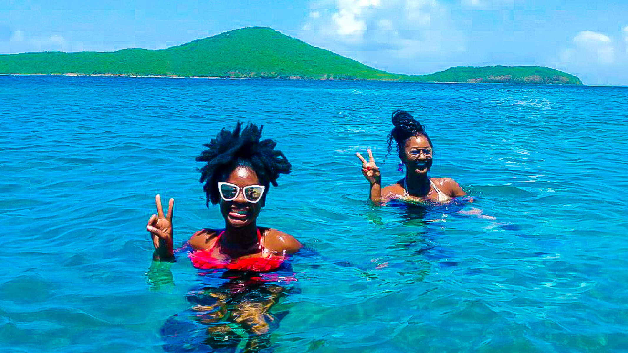 two melanated women throwing up peace signs in the crystal blue waters around Culebra, Puerto Rico, on a day trip.