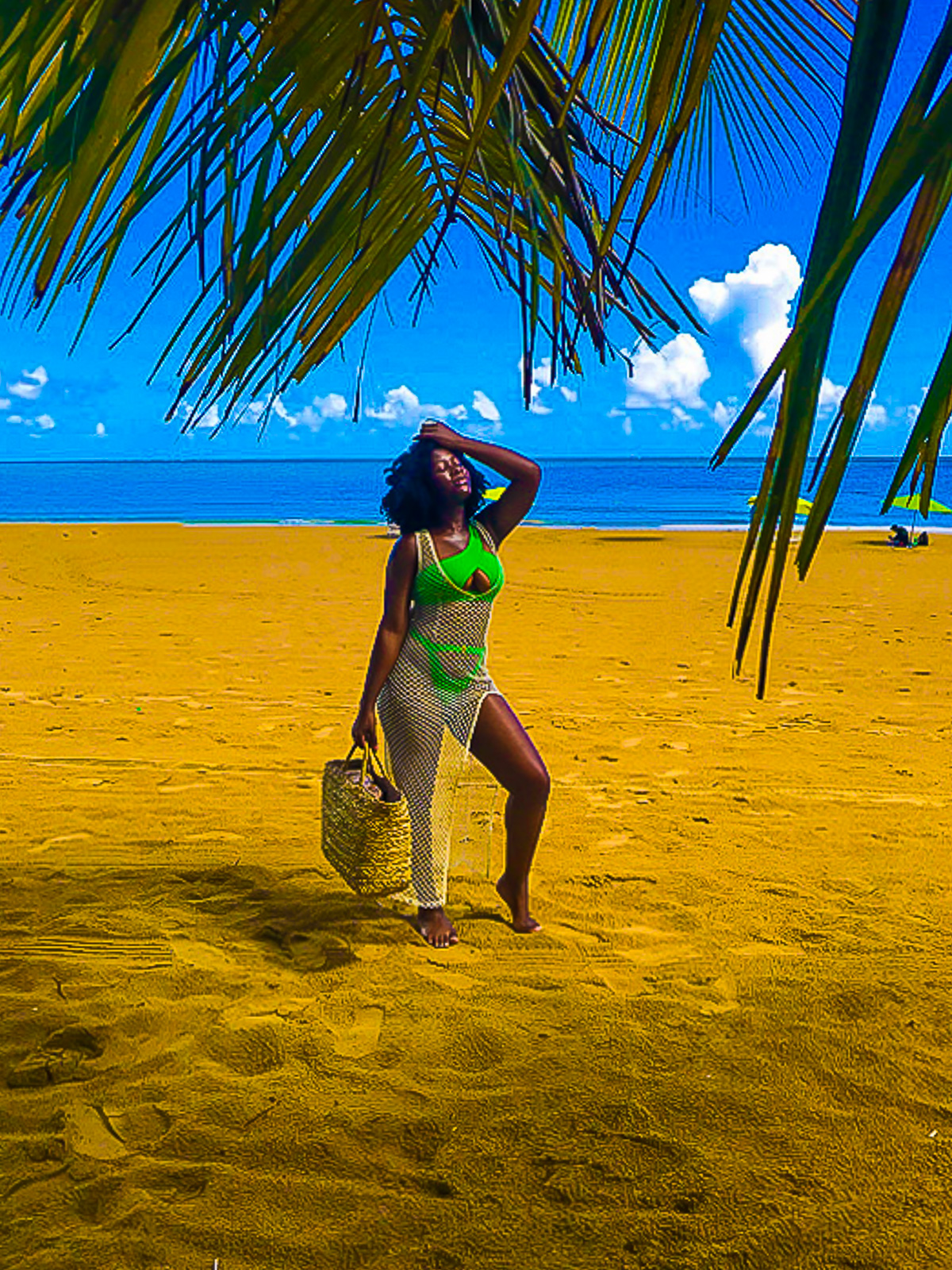 Black girl posing on Isla Verde beach in San Juan, Puerto Rico, wearing a bright green bikini with a mesh cover up dress and straw beach bag.