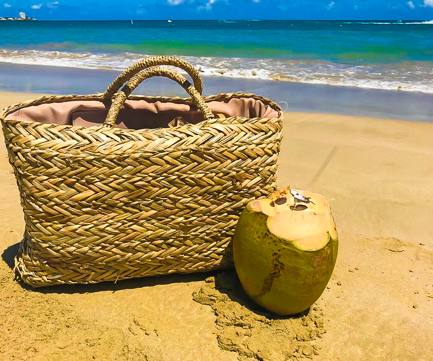 large straw beach bag next to a drinking coconut in the sand on Playa Isla Verde, PR.