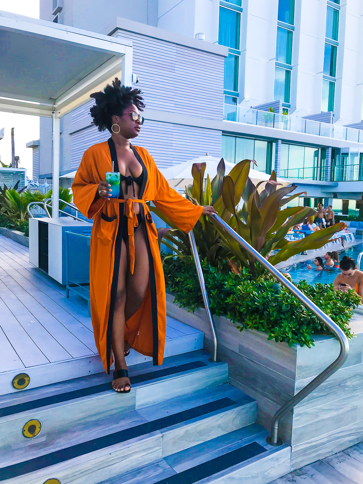 Jazzmine at Serafina Beach Resort cabana wearing an orange and black belted kimono cover up and holding a mojito cocktail.