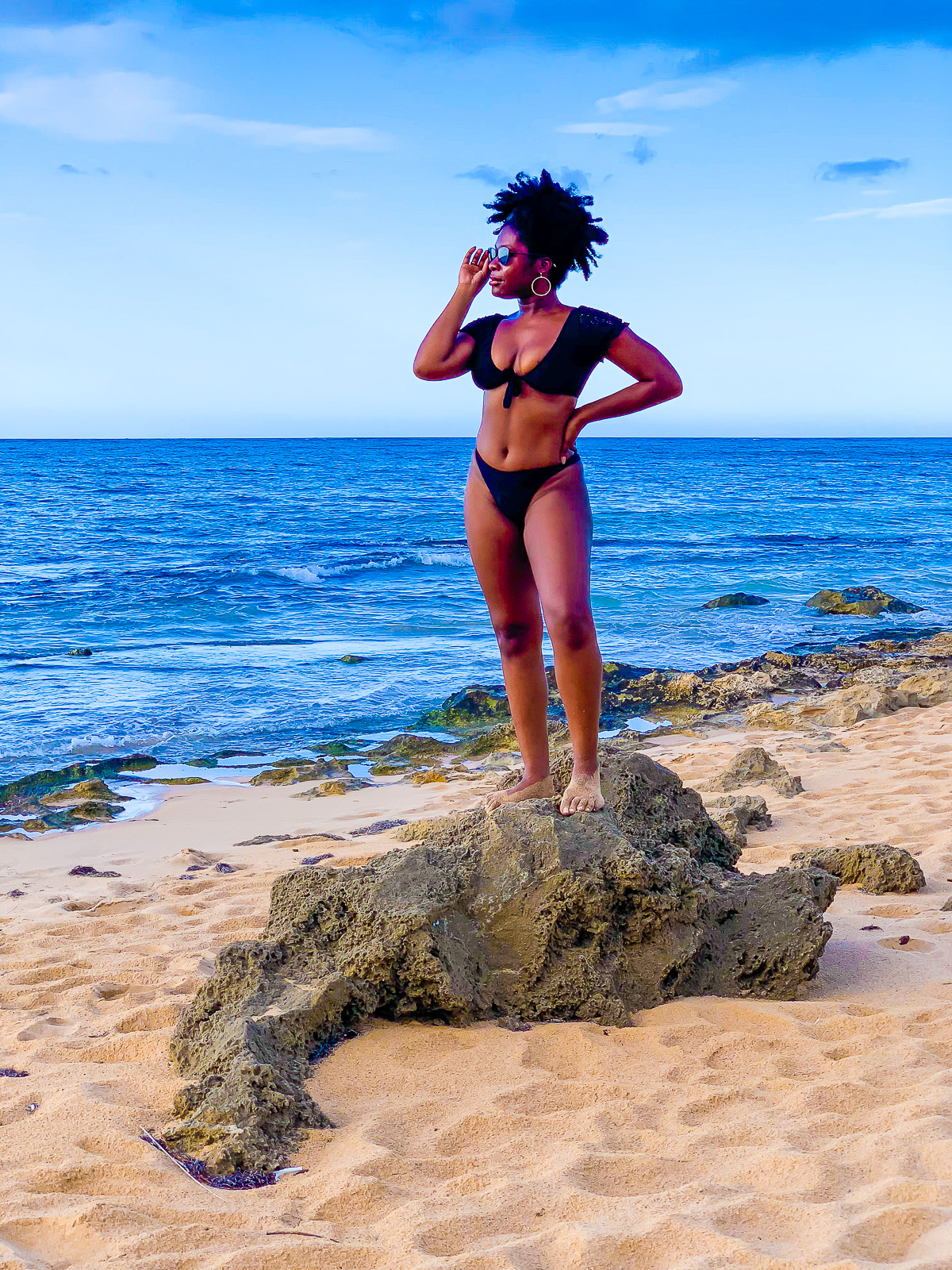Jazzmine standing on coral rock on Condado Beach wearing a black crochet bikini and gold hoop earrings.