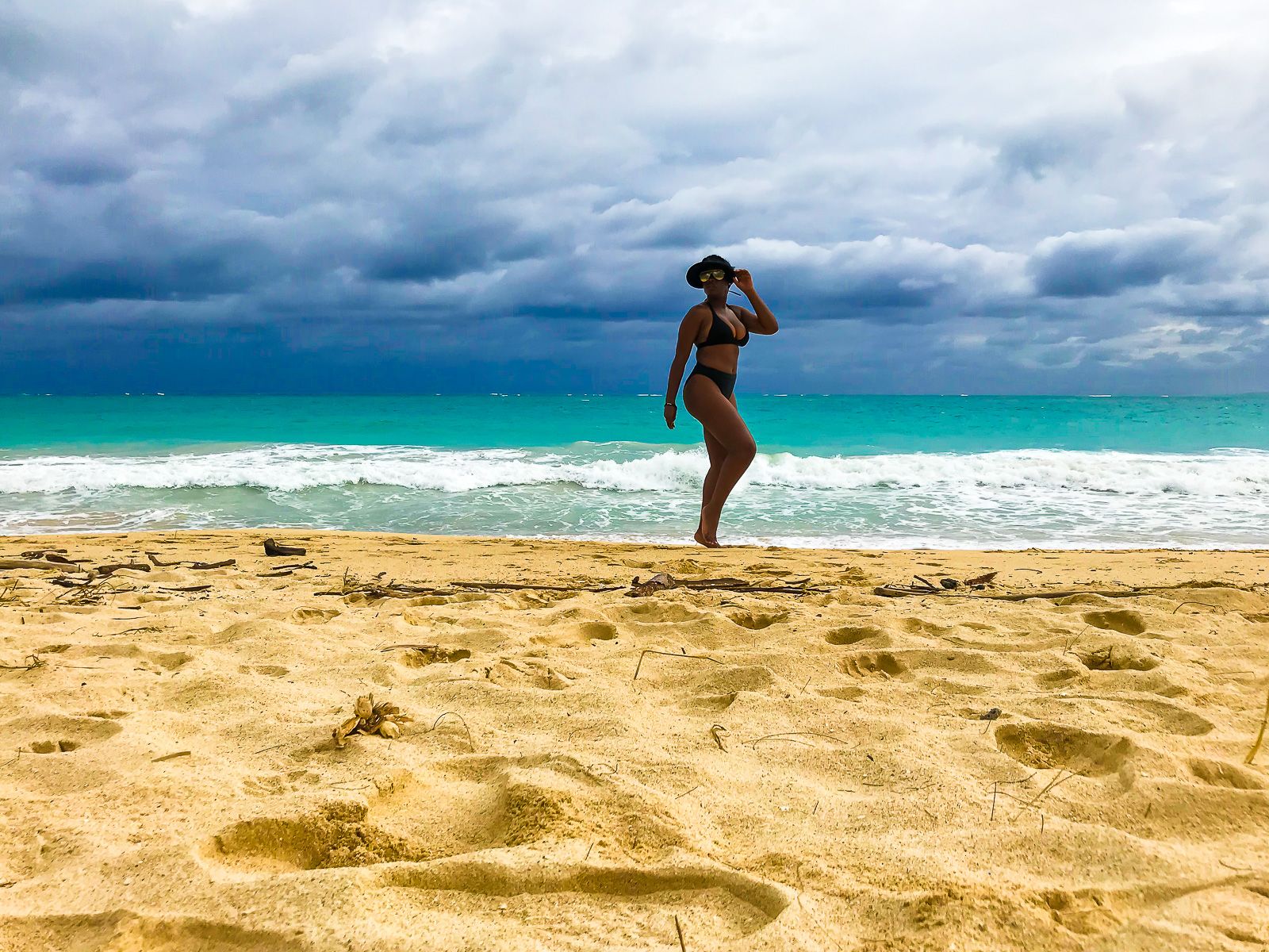Jazzmine walking along the water at Bellow Airfield Beach Oahu, Hawaii, wearing high-waist black bikini and visor.
