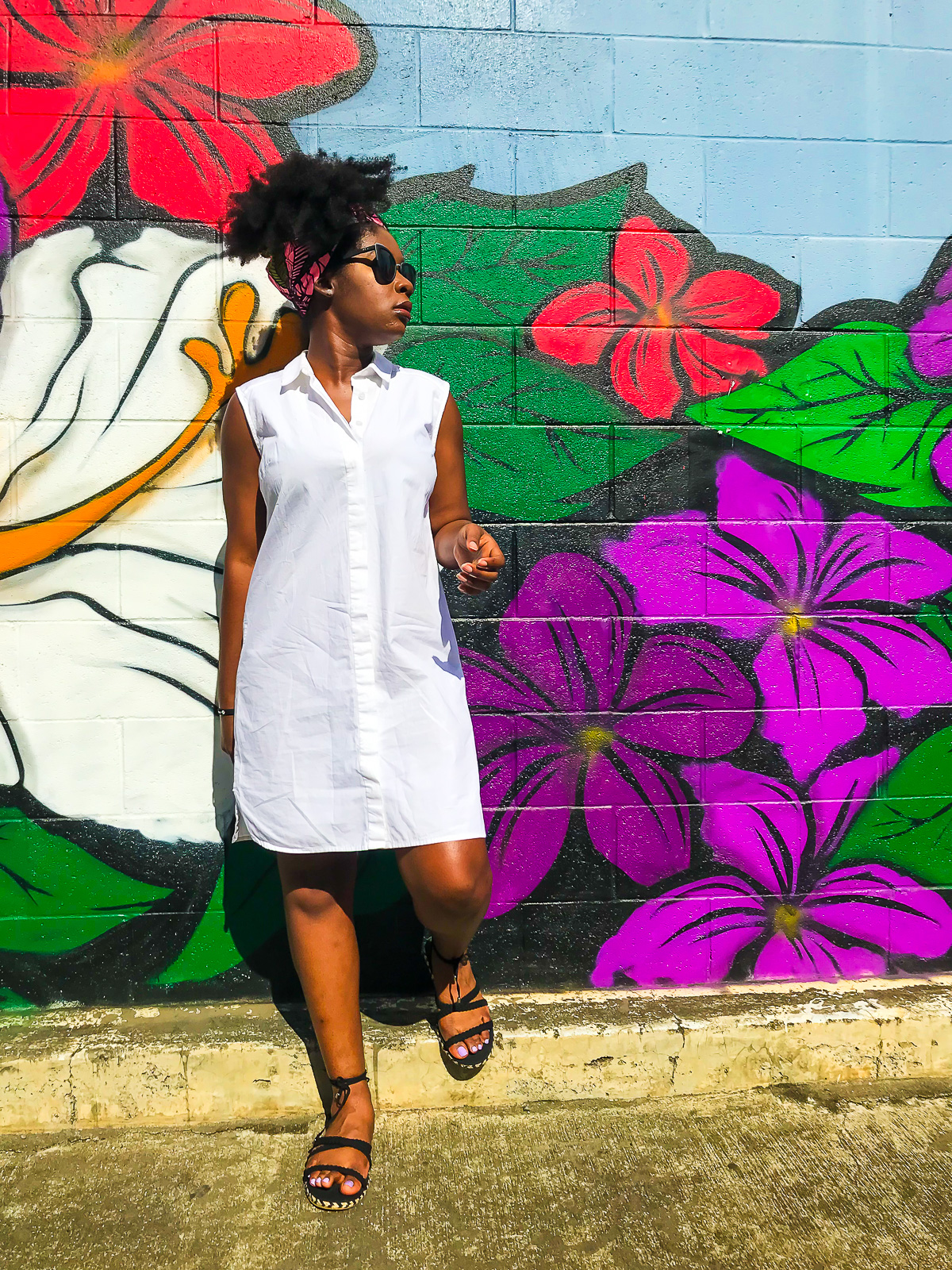 Jazzmine standing in front of a vibrant purple and red street mural in Kaka'ako, wearing a crisp white sleeveless shirt dress, pink and green head wrap, and black espadrilles.
