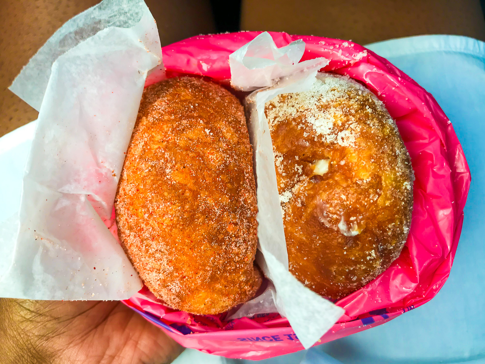 brown hand holding two malasadas wrapped in hot pink bag from Leonard's in Oahu, Hawaii.