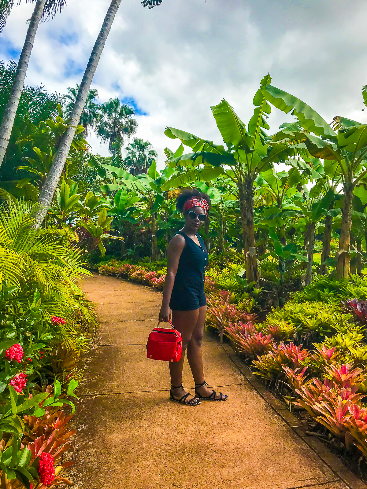 Jazzmine wearing a black linen romper, black sandals, and red hair tie with a red bag standing in a walkway at Oahu's Dole Plantation.