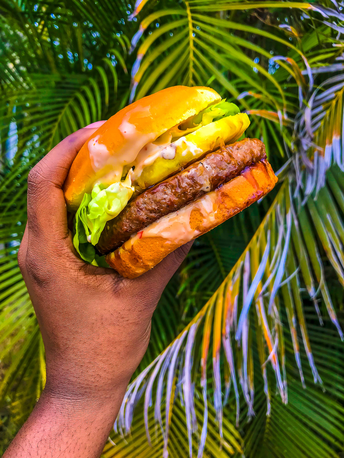 brown hand holding juicy Kualoa Ranch burger in front of palm fronds.