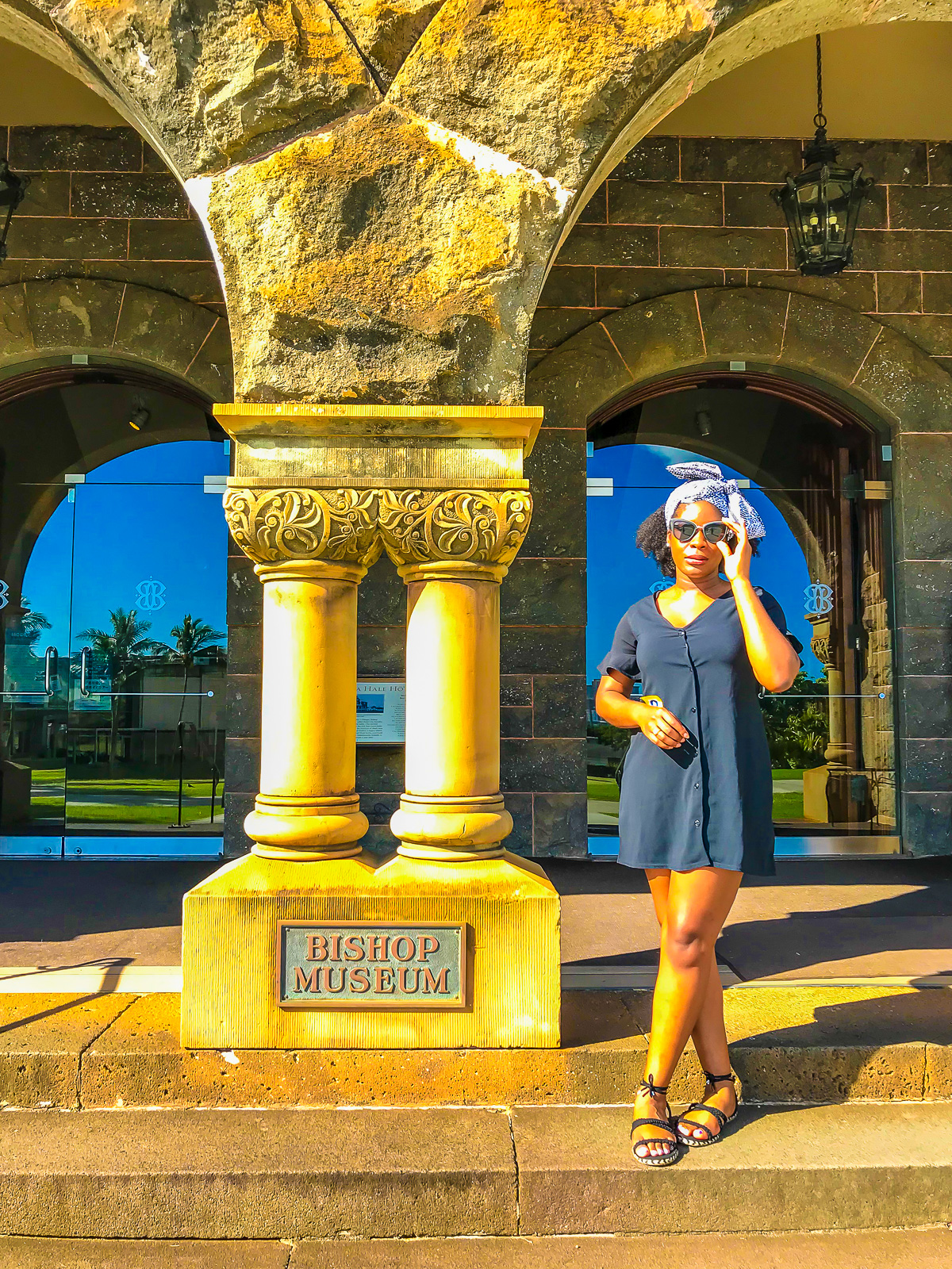 young woman posing outside Bishop Musuem on Oahu wearing a black button-down mini dress, lace up sandals, and black and white ankara head scarf.