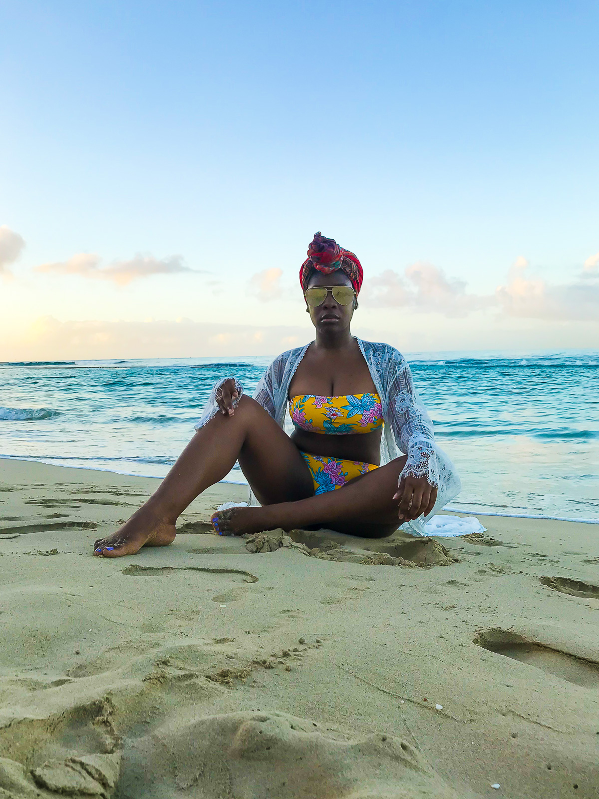 Black woman sitting in the sand on Waikiki shore wearing a yellow floral bikini, white lace kimono cover-up and gold polarized aviators.