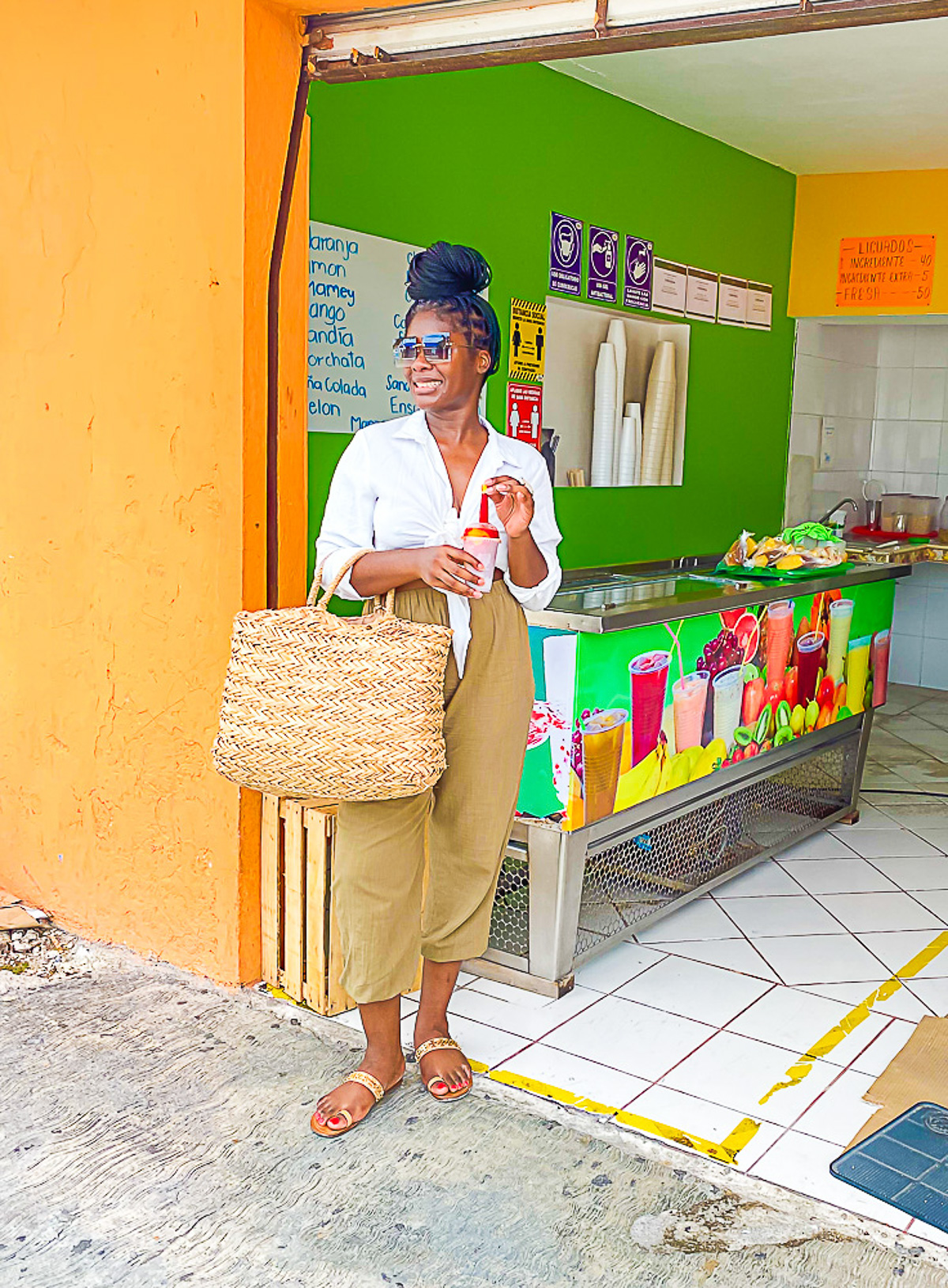 young woman dressed for vacation in Cozumel wearing green linen pants, sandals, a white cropped top, and a straw beach bag.