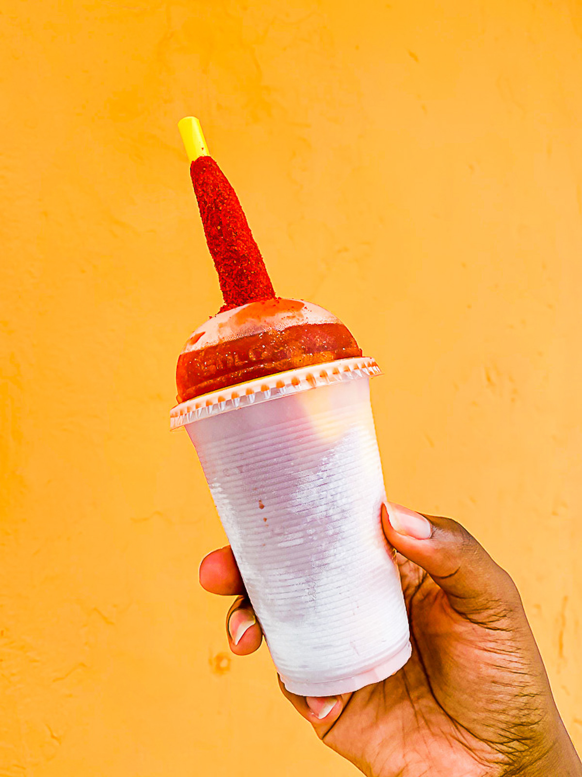 brown hand holding a mango chamoyada in a frosty plastic cup in front of a bright orange paint wall in Cozumel, Mexico.