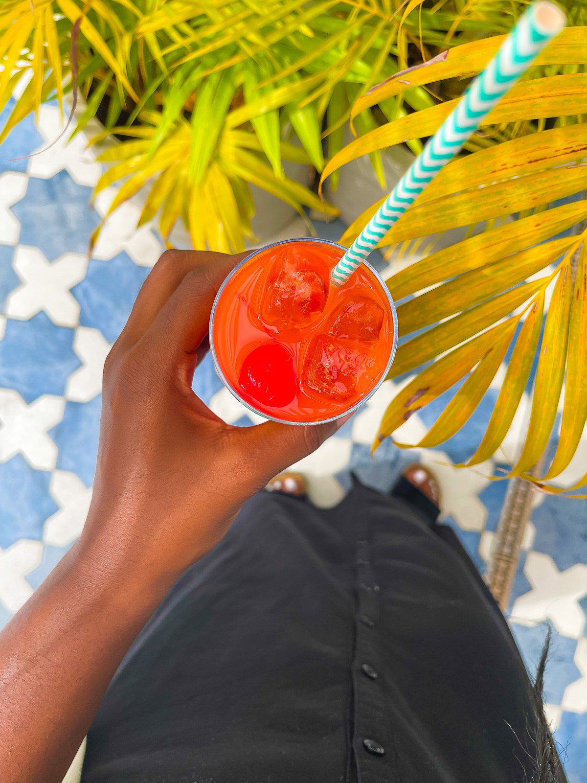 brown hand holding a bright red welcome mocktail with a striped paper straw at a West Indian resort in Grenada.