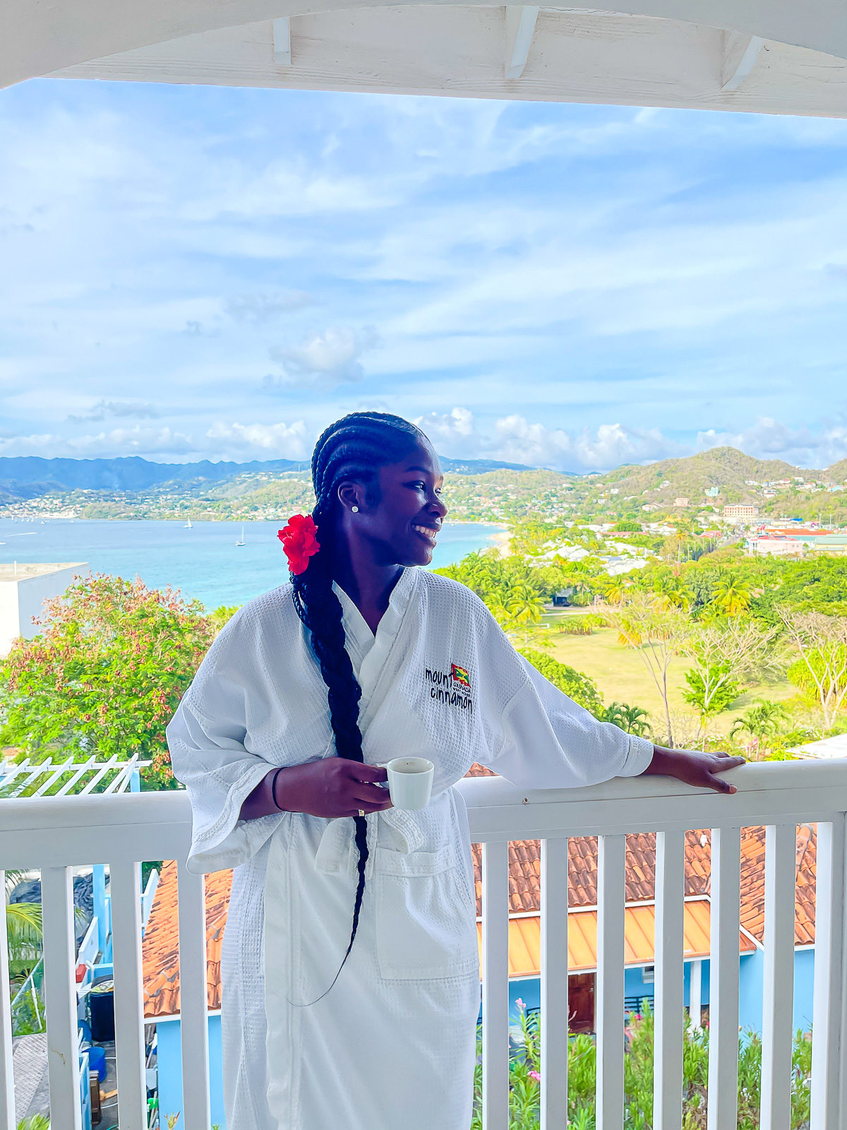 Jazzmine standing on room balcony wearing a spa robe and red flower in her hair at Mount Cinnamon Resort with Grand Anse Beach in the background.