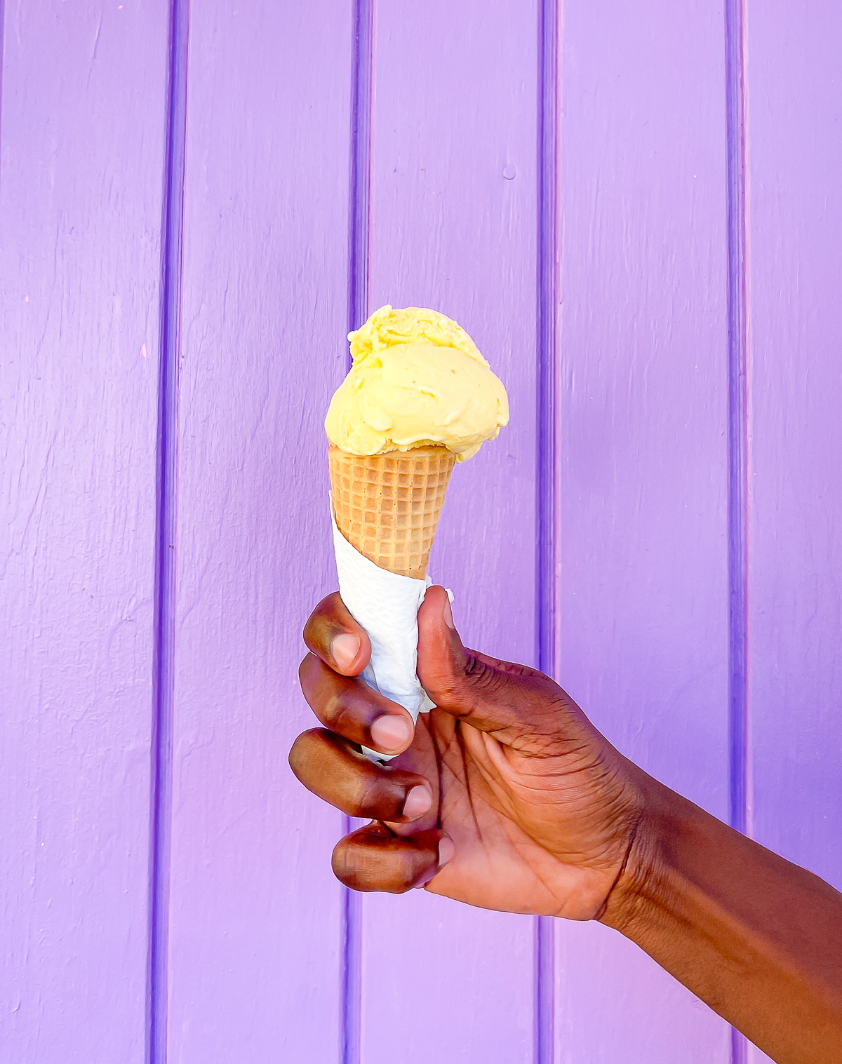 brown hand holding waffle cone piled with Grenadian nutmeg ice cream in front of a purple painted wall.
