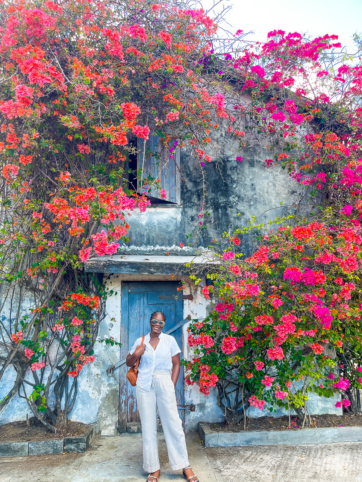 Jazzmine standing in front of a wall covered in Bougainvillea, wearing white cotton camp shirt and flax linen pants in Tivoli, Grenada.
