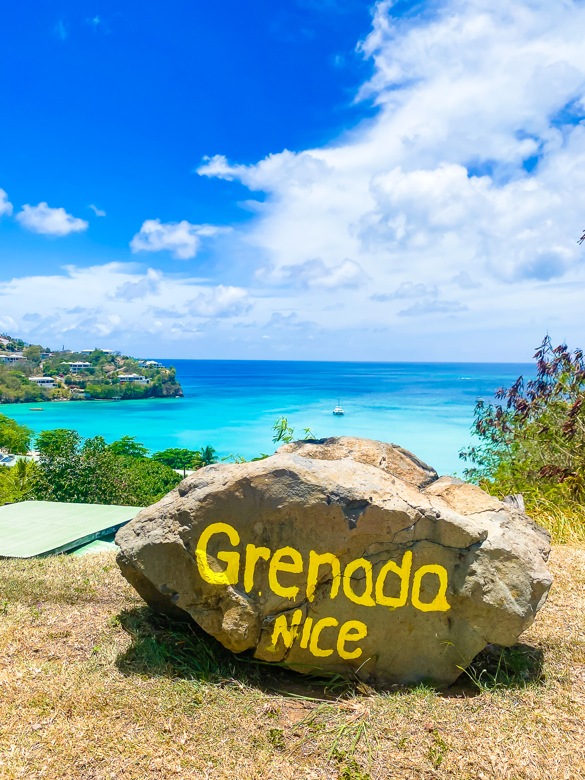 large stone with the words "Grenada Nice" painted on it in yellow; Morne Rouge Bay flowing out into the Caribbean Sea behind it.