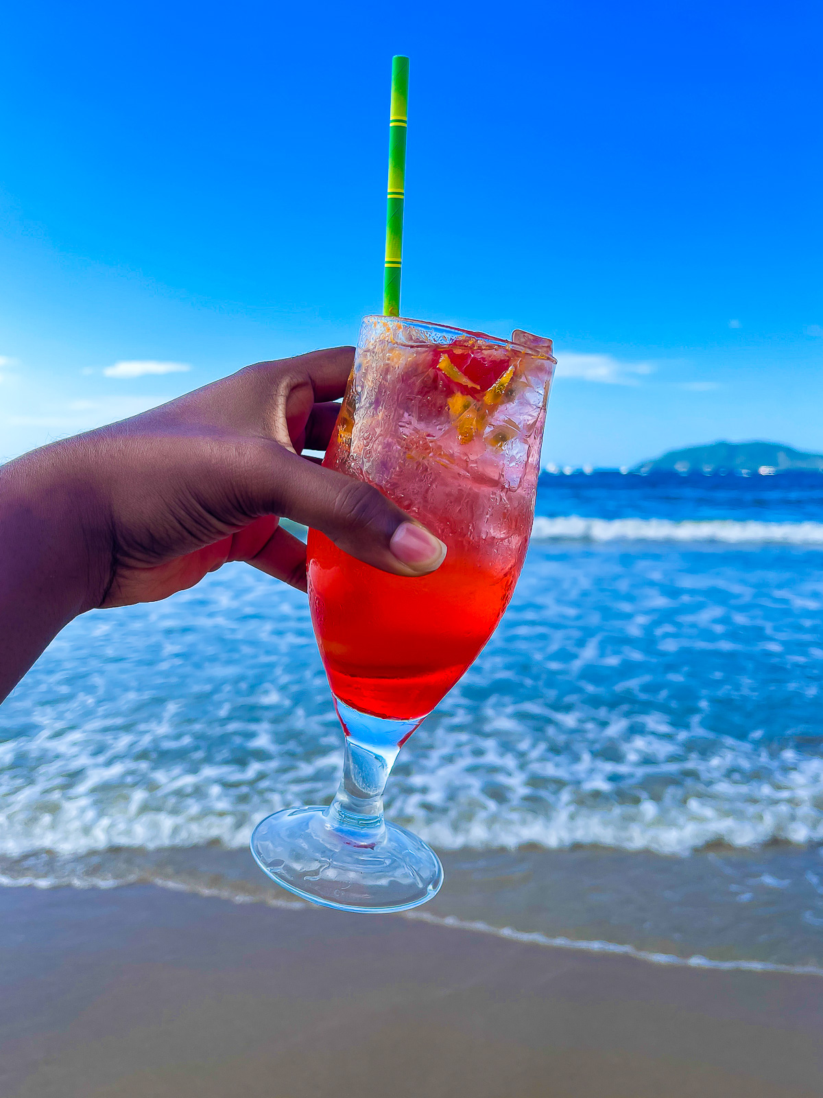 brown hand holding up a colorful red cocktail with a bright green straw in from of the receding tide on Tamarindo Beach in Costa Rica, Central America.